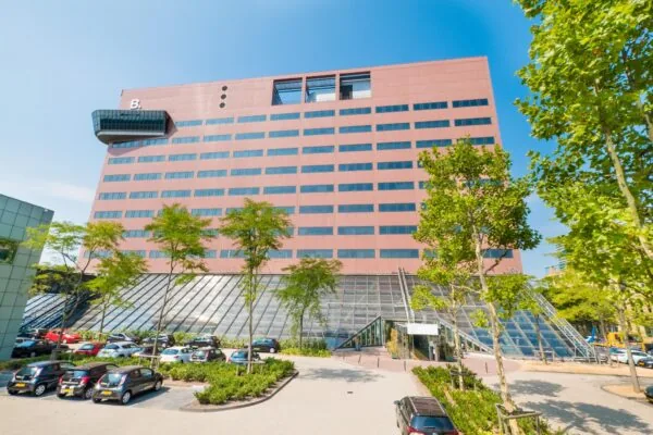 Modern kantoorgebouw aan de Johan Huizingalaan in Amsterdam met parkeerplaatsen en glazen entree, gezien op een zonnige dag.